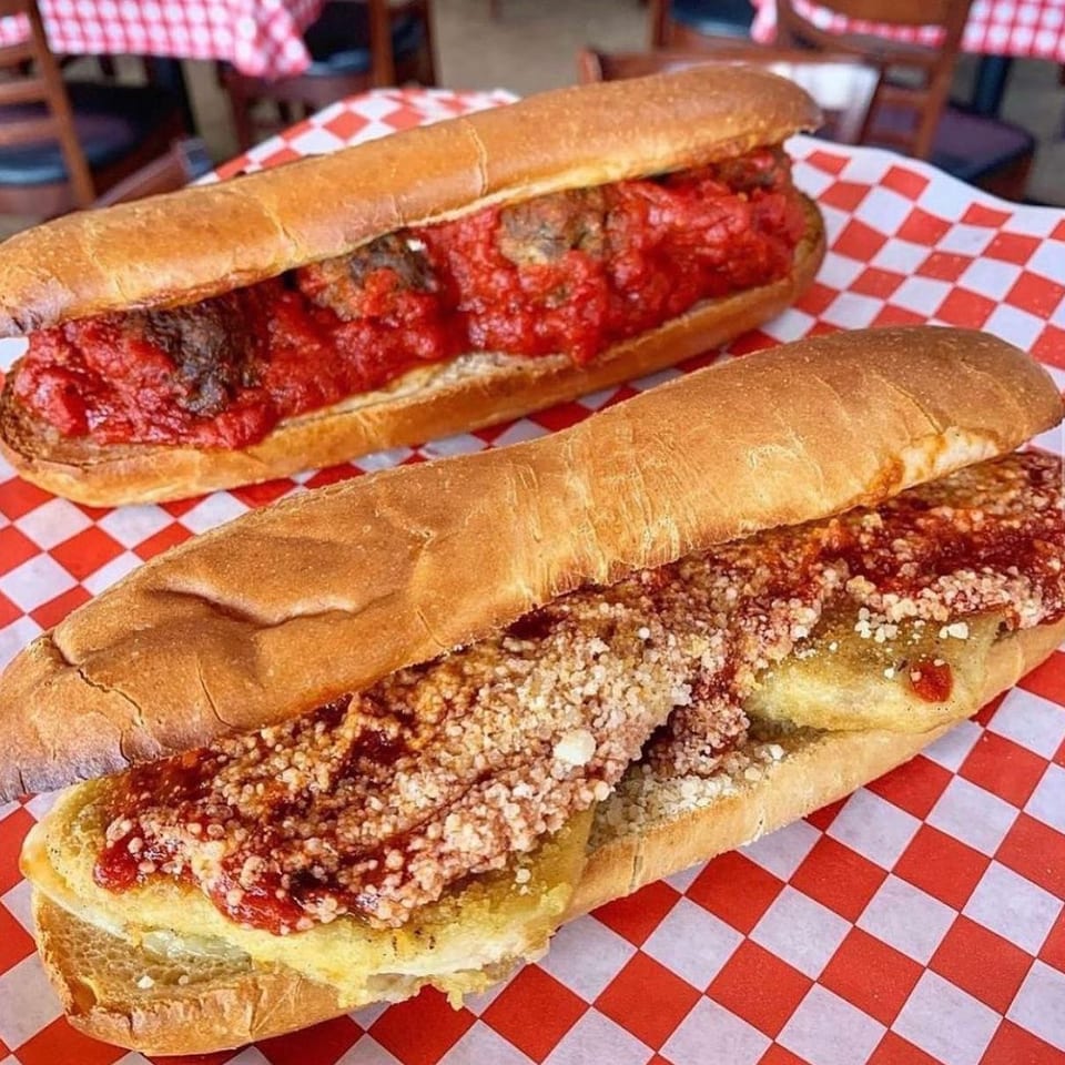 Two saucy meatball subs on crusty rolls, one topped with grated cheese, served on red-and-white checkered paper.