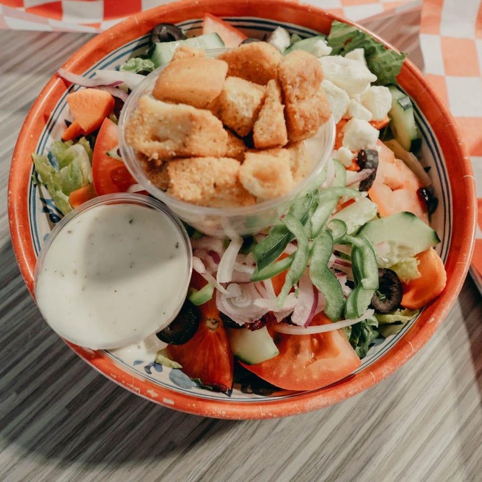 Garden salad topped with crispy chicken pieces, ranch dressing, and vegetables beside a bowl of spaghetti on checkered paper.
