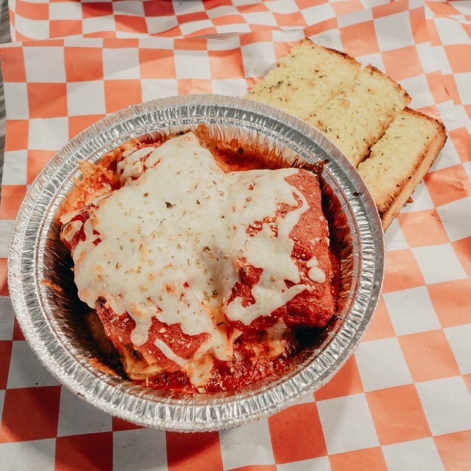 Cheesy baked pasta in marinara sauce served in a foil dish with garlic bread on orange-and-white checkered paper.