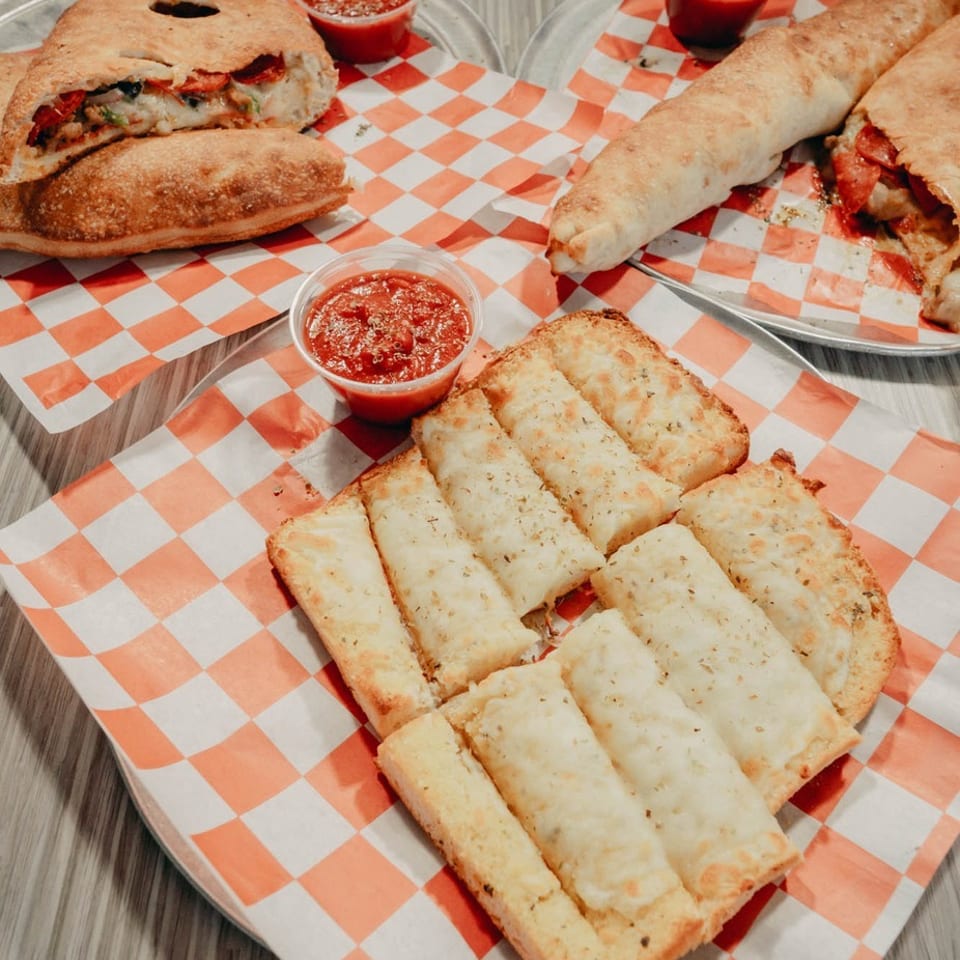 Assorted Italian sandwiches, breadsticks, and marinara dipping cups served on orange-and-white checkered trays.