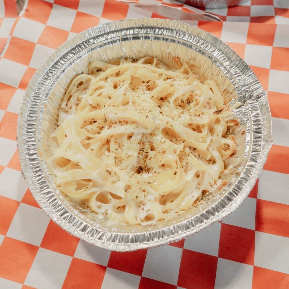 Alfredo pasta topped with grated cheese in a foil tray, with cheesy baked pasta behind it on red-and-white checkered paper.