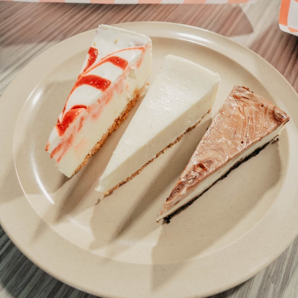 Three cheesecake slices, including raspberry-swirl, plain, and marbled chocolate varieties, arranged on a plate beside a bread roll.