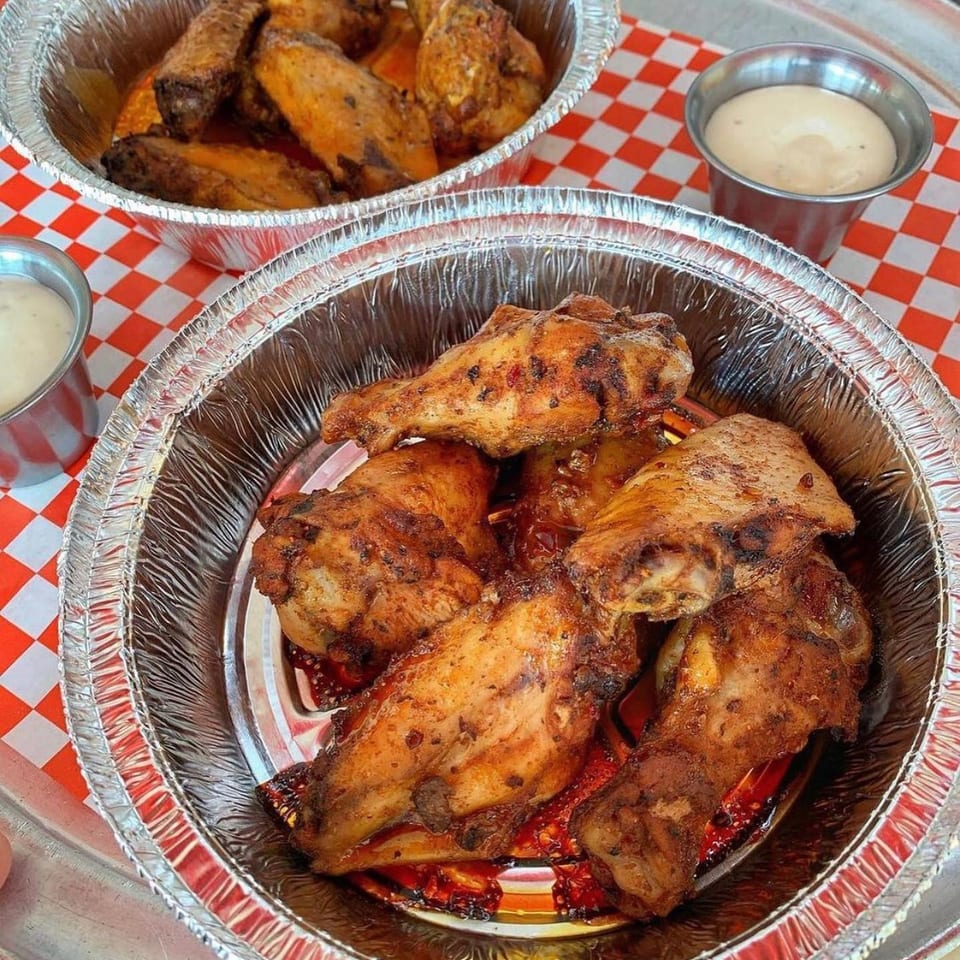 Grilled seasoned chicken pieces in foil trays with creamy dipping sauce on a red-and-white checkered tablecloth.